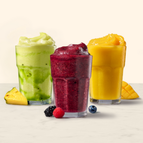 Three frappes, one green, one purple and one yellow, with fruit surrounding the frappes.